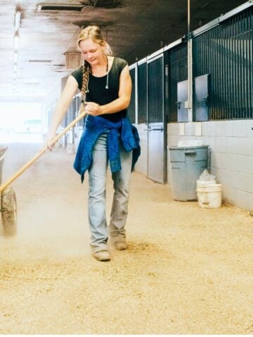 Woman raking tidy horse barn aisle to control dust and improve horse barn air quality.